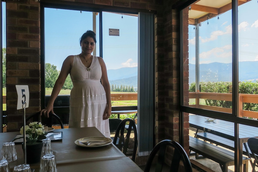 Geetu stands by a table set up for dining at her restaurant, out the window are rolling mountains in a blue sky.