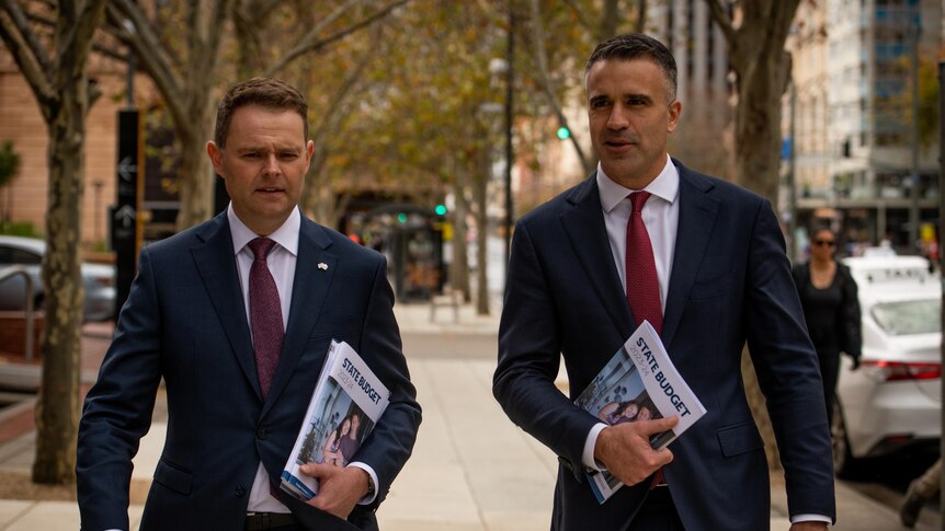 Two men walk down a city street holding budget papers in their hands