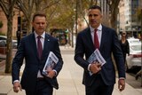 Two men walk down a city street holding budget papers in their hands