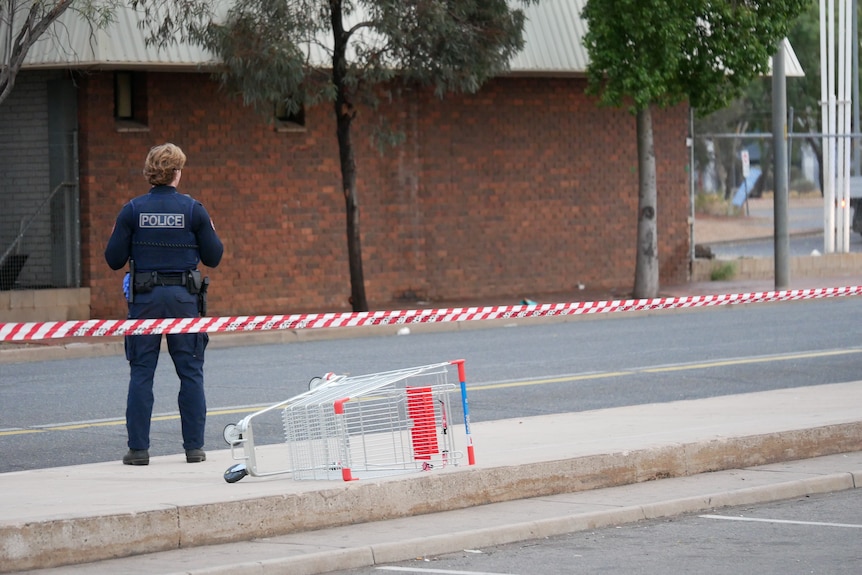 A police officer standing next to an overturned shopping trolley with police tape cordoning off an area.