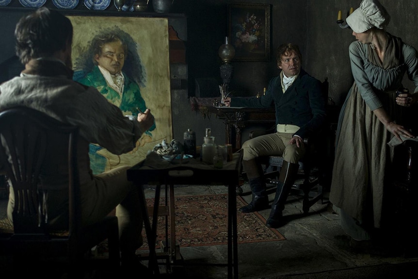 Colour still of Ben Crompton painting seated Rory Kinnear, who is also talking to Bryony Miller in 2018 film Peterloo.