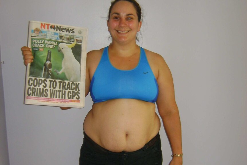 A woman with a chubby belly in a crop top stands with a picture of a newspaper for a "before" weight loss photo.