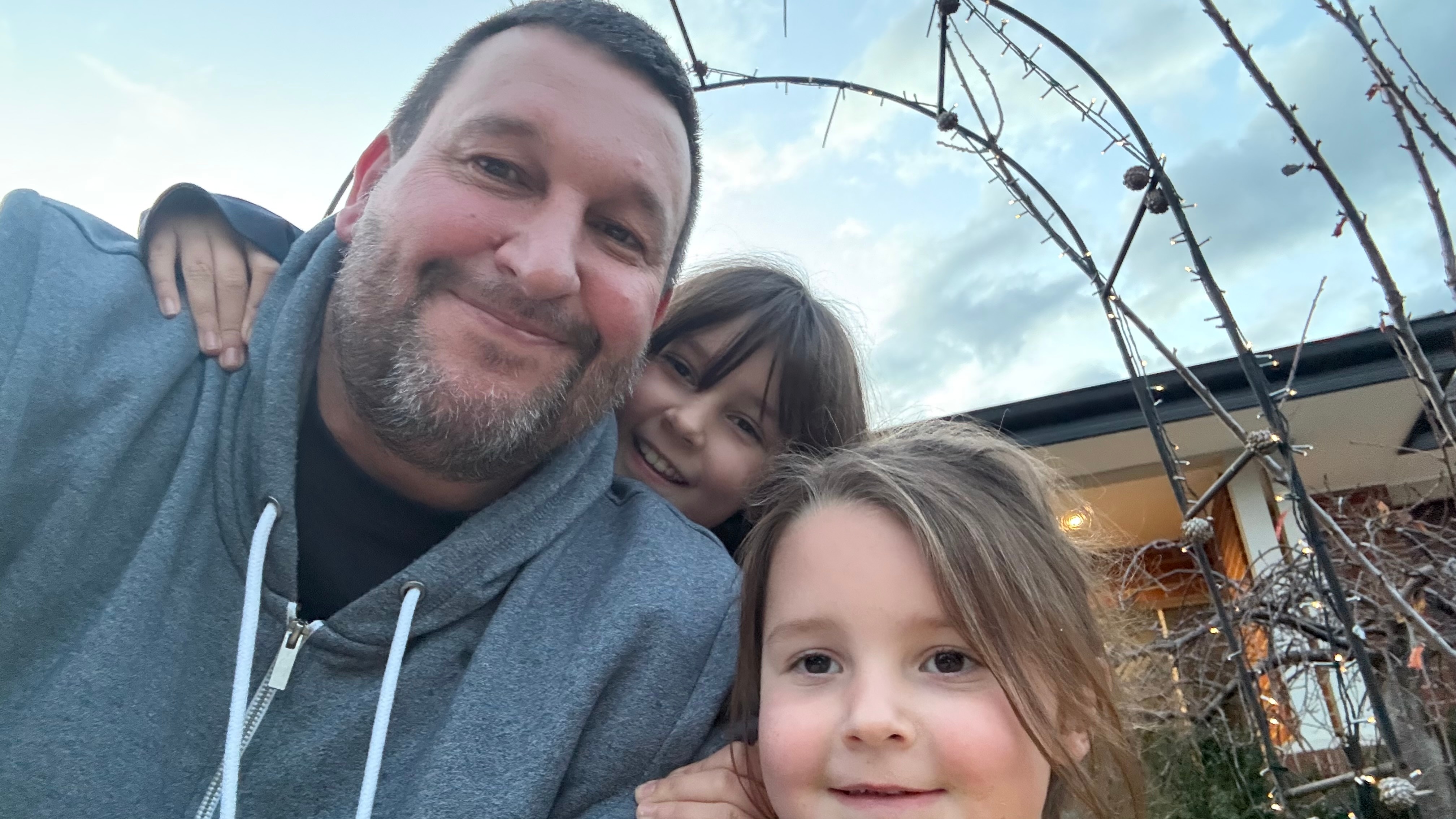 selfie of Steve with his two children, blue sky behind, a trellis with some wires.