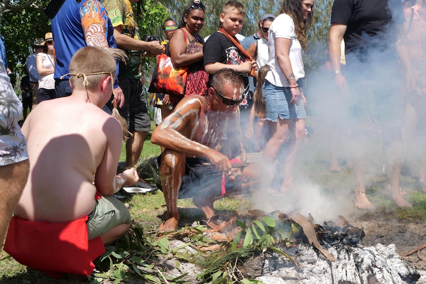A painted Indigenous man crouches by a smoking ceremony while people walk around him