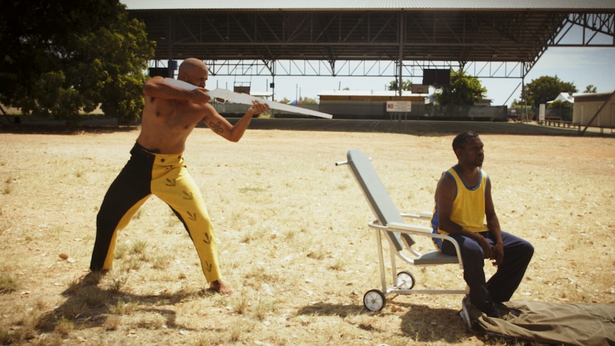 A shirtless Aboriginal man with a white gun aimed at an Aboriginal man sitting on a white chair