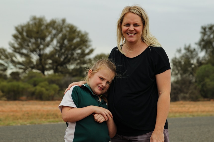 A woman stands roadside with her arm around her young daughter, smiling at the camera.