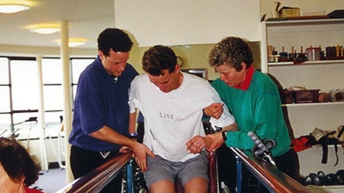 A man is helped out of a wheelchair by two people as he learns to walk again in physical therapy ward following an accident.