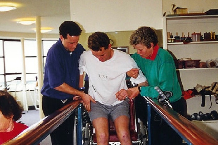 A man is helped out of a wheelchair by two people as he learns to walk again in physical therapy ward following an accident.