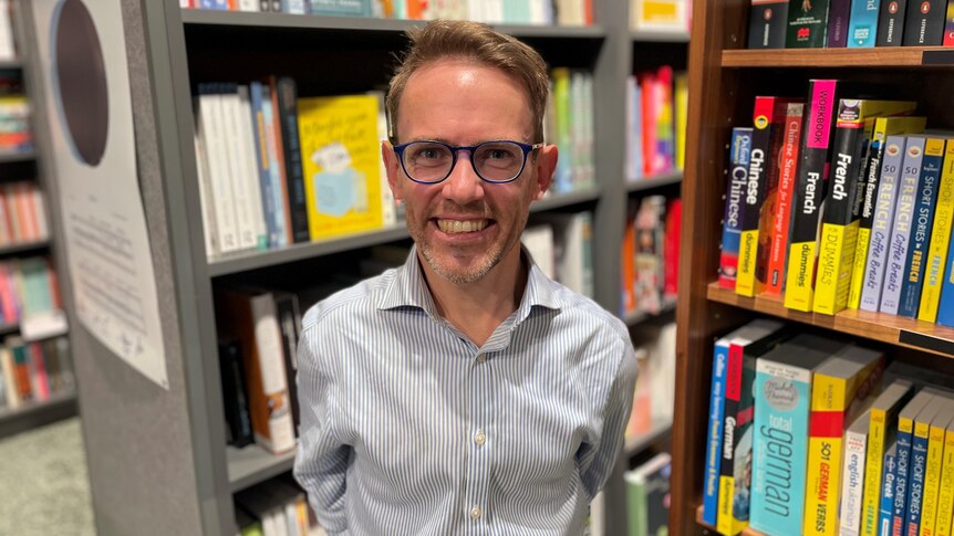 Sam Drummond is smiling. He is wearing glasses and a blue button up shirt, and is standing in front of bookshelves.