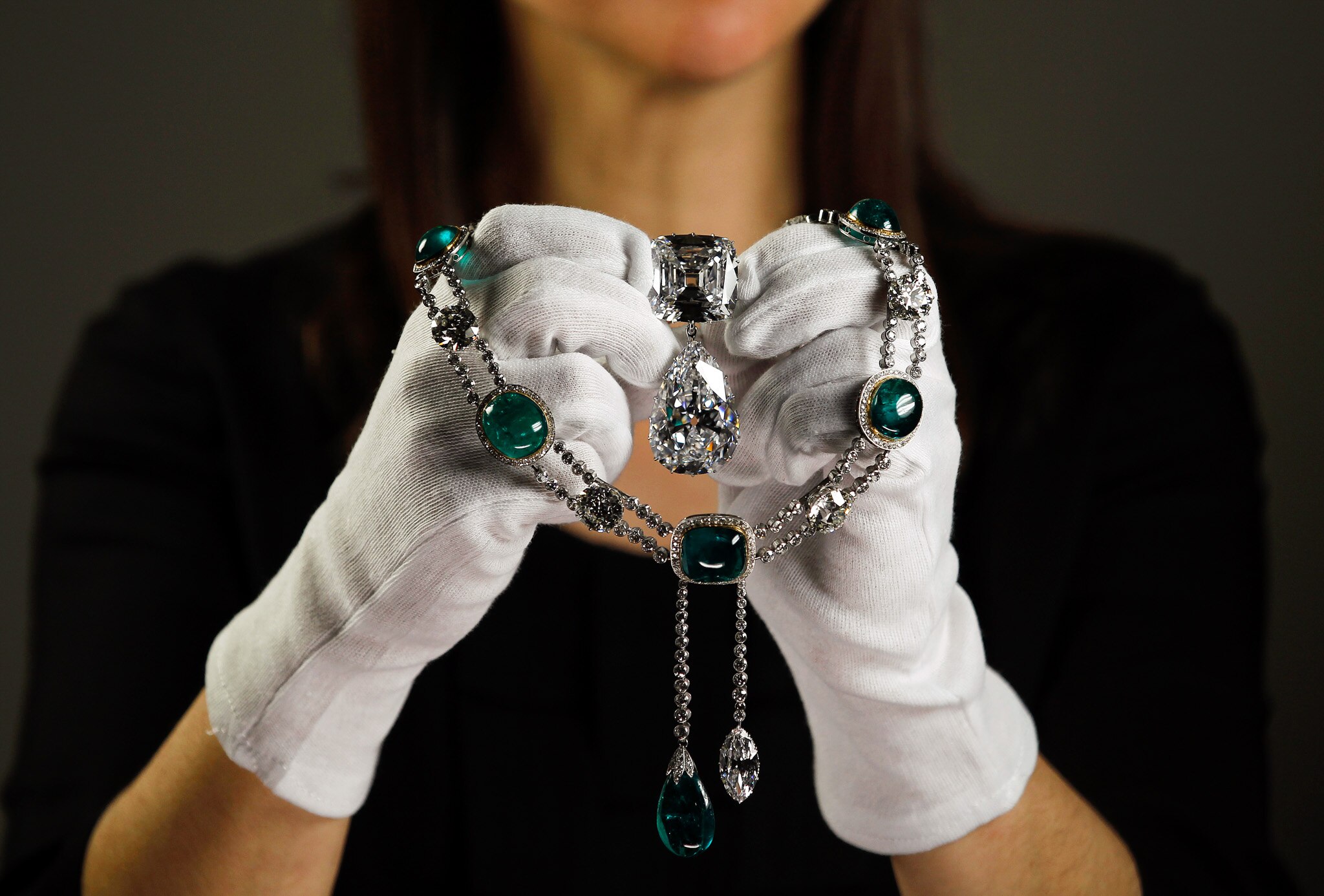A woman with white gloves holds up a  necklace with emeralds and a big diamond in the centre.