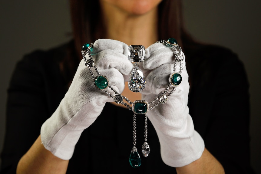A woman with white gloves holds up a  necklace with emeralds and a big diamond in the centre.