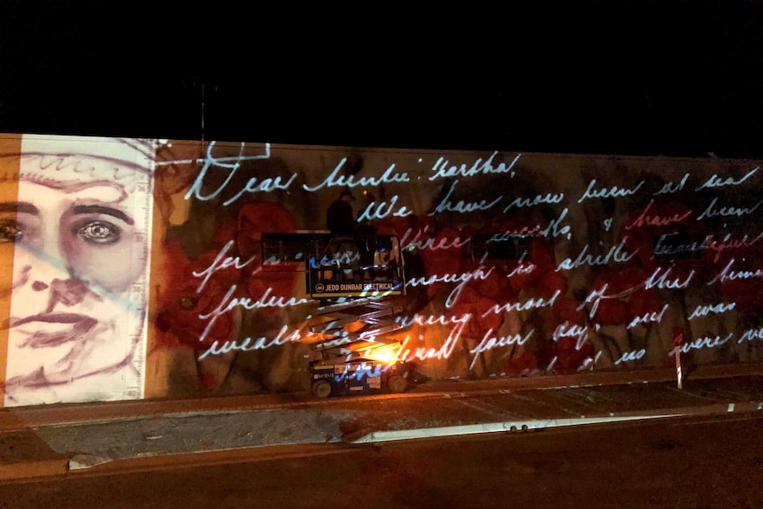 A spray painter stands on a cherry picker at night while tracing handwriting projected on a wall with a movie projector.