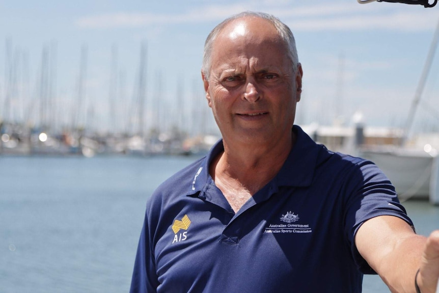 Peter stands near a wharf smiling in front of sail boats, wearing an AIS t-shirt.