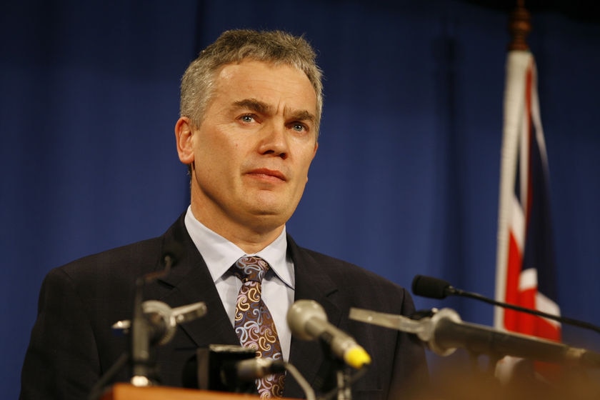 John Lenders, wearing a suit and tie, stands before several microphones at a lectern.