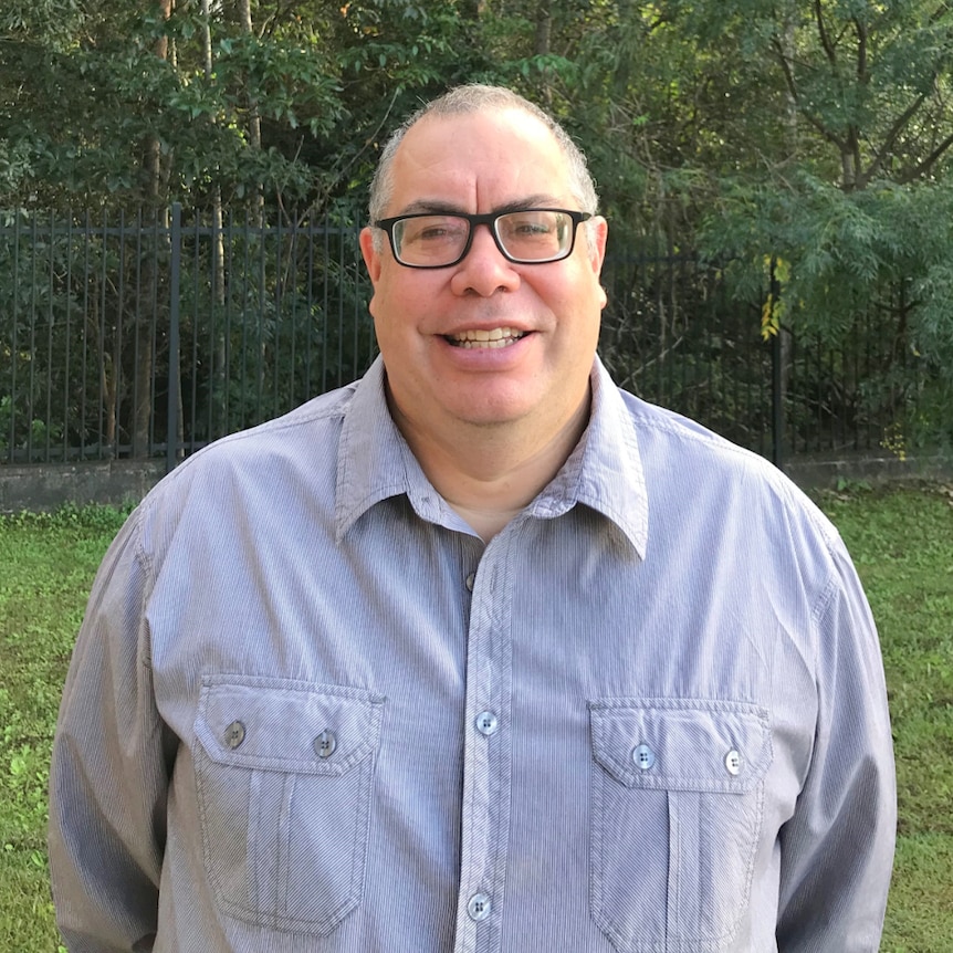 Man in button up shirt smiling with green grass and trees in background