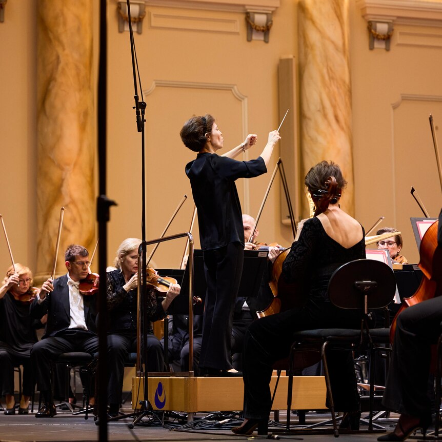 Teresa Riveiro Böhm wearing black, stands on a podum conducting an orchestra.