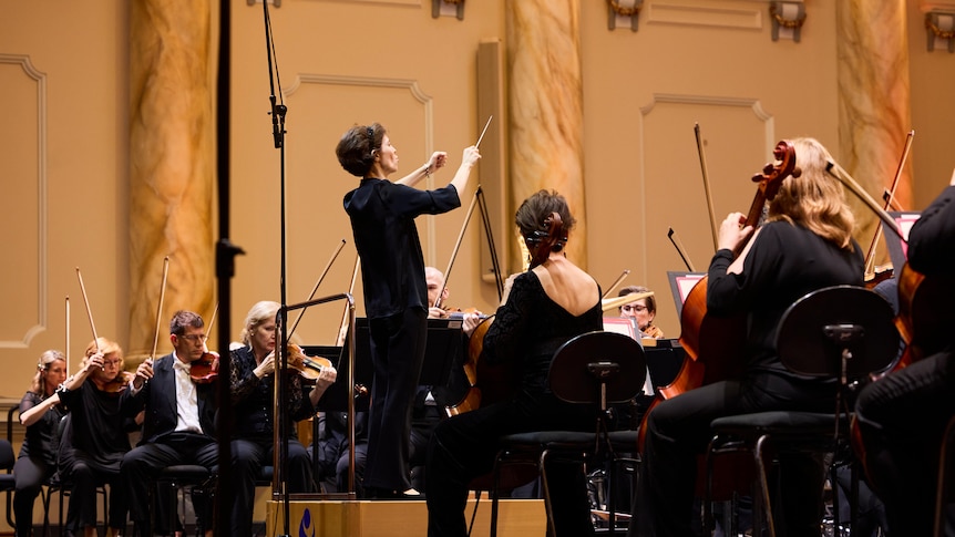 Teresa Riveiro Böhm wearing black, stands on a podum conducting an orchestra.