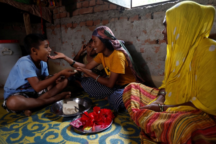 Three people, two women and a young boy, sit on the flood with a tray of hibiscus flowers and cake between them. 