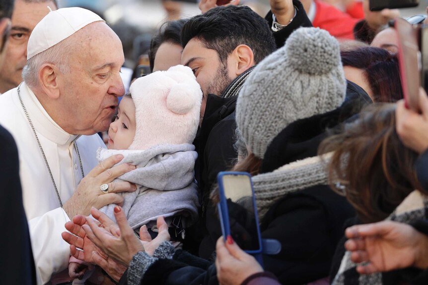 The Pope kisses a baby dressed in winter clothes in a crowd of people.