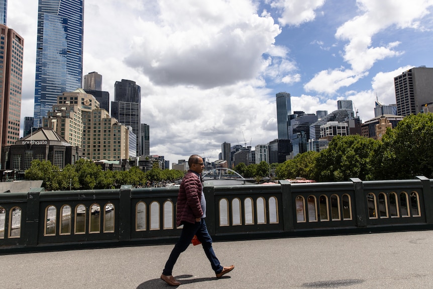 A man wearing a puffy jacket walks across a bridge with tall buildings behind