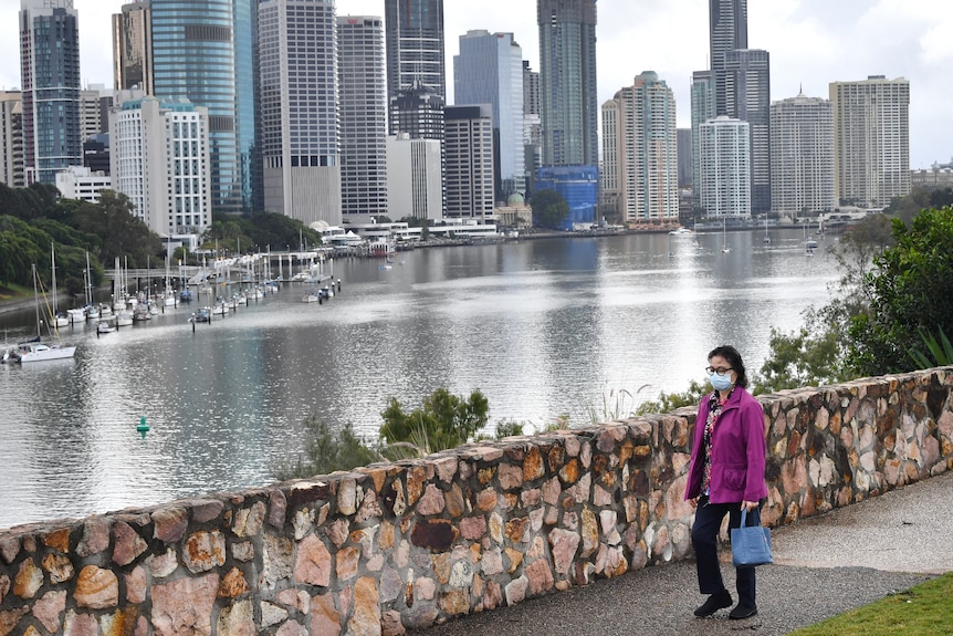 A woman wears a face mask walking along the Kangaroo Point Cliffs with city buildings in view in Brisbane