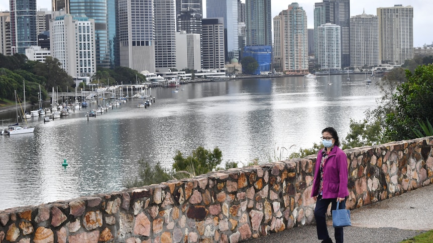 A woman wears a face mask walking along the Kangaroo Point Cliffs with city buildings in view in Brisbane