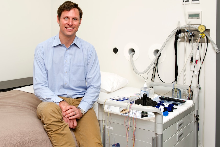 A man wearing a blue shirt and brown pants sits on side of bed next to wires and a machine used in sleep studies.