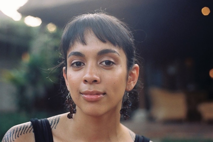 Une femme sourit en regardant directement la caméra, elle a des boucles d'oreilles noires pendantes.