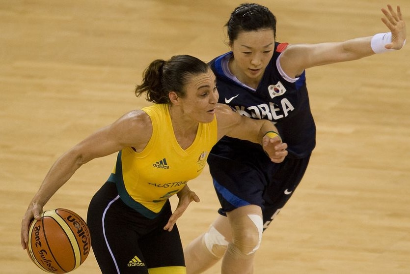 A woman with brown hair in a pony tail wearing an Australian basketball uniform fends off a Korean opponent 