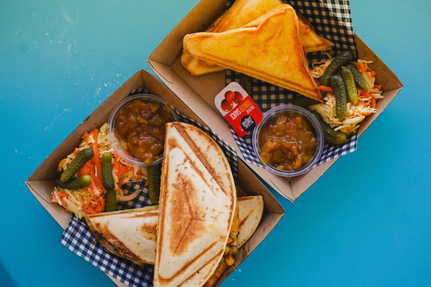 Two cardboard food trays lined with checkered paper with a small slaw, toasted sandwich and relish on each.