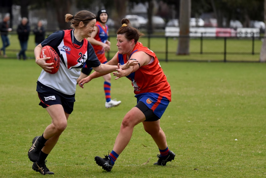 A woman holds a Sherrin and fends off a player who is trying to tackle her.