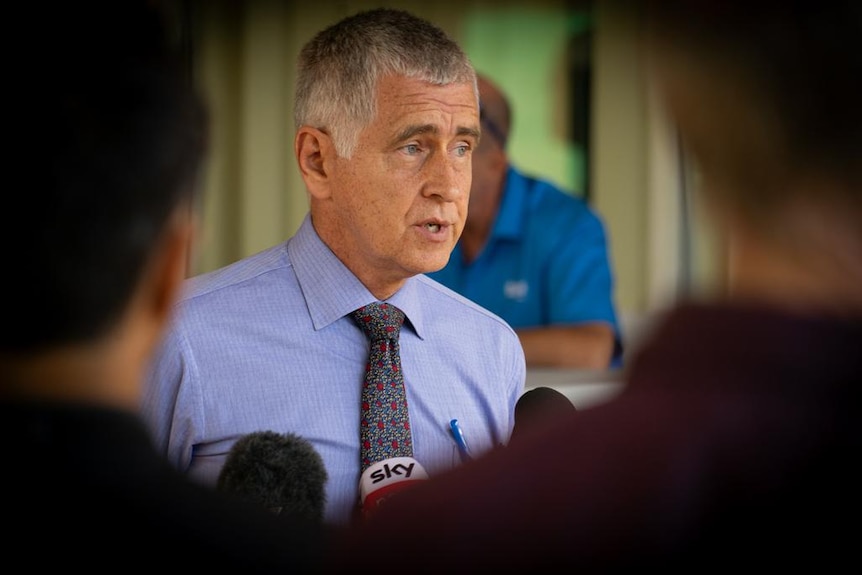 A middle-aged man wears a collared shirt and tie, at a press conference