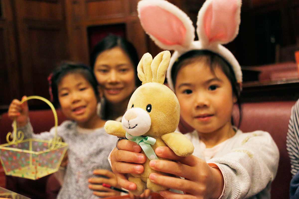 A little girl wearing rabbit ears holds out a toy Easter bunny.