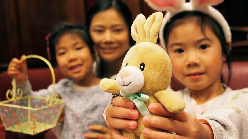 A little girl wearing rabbit ears holds out a toy Easter bunny.