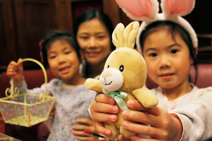 A little girl wearing rabbit ears holds out a toy Easter bunny.
