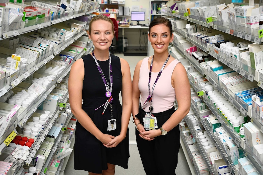 Two young pharmacy graduates stand in between two aisles of pharmacy products.