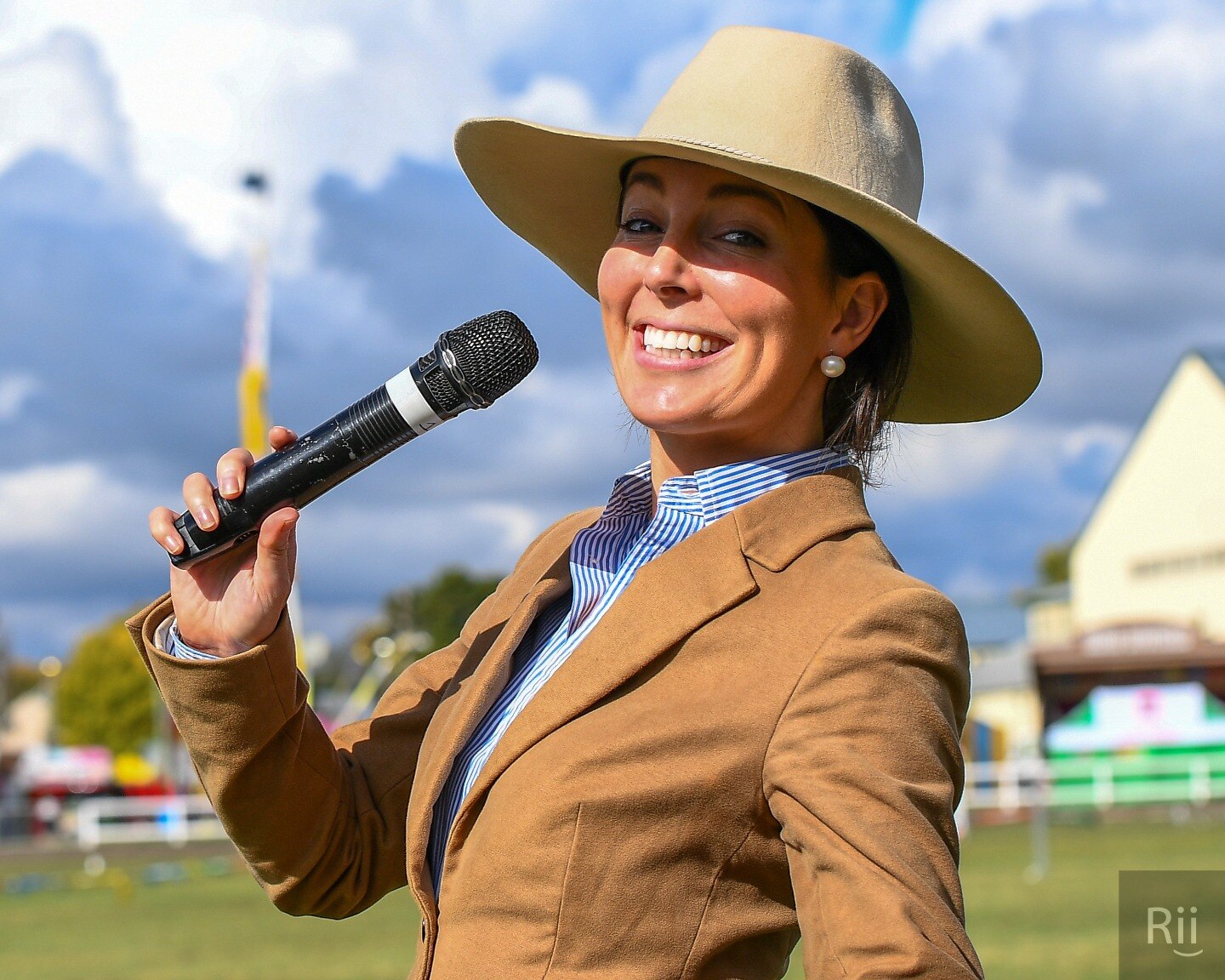 A young woman smiles while holding a mic to her mouth. She's dressed in a fitted blazer wearing an akubra
