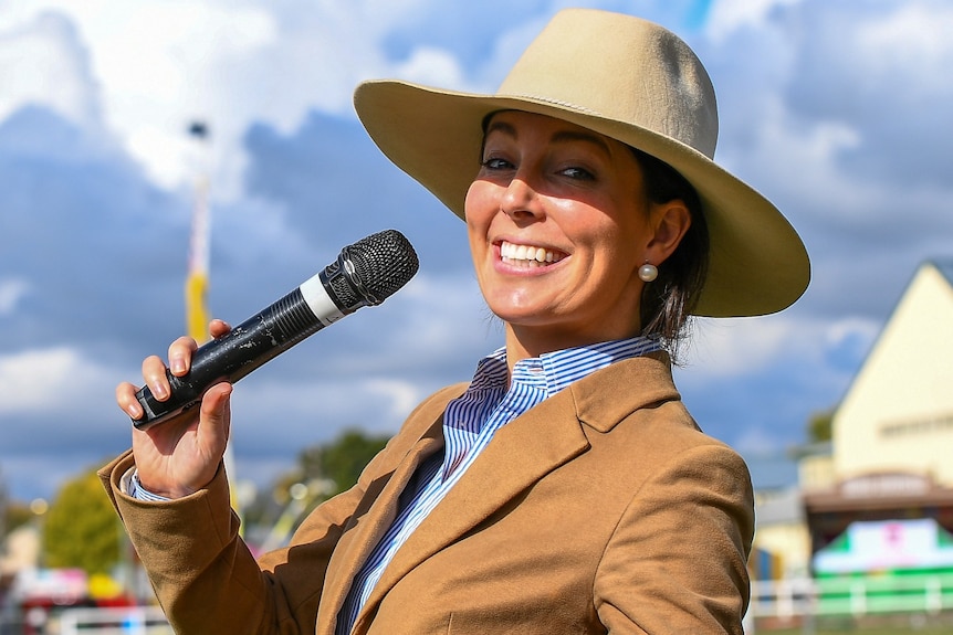 A young woman smiles while holding a mic to her mouth. She's dressed in a fitted blazer wearing an akubra