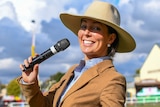 A young woman smiles while holding a mic to her mouth. She's dressed in a fitted blazer wearing an akubra