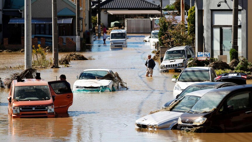 Partially submerged cars are seen in a suburban street, with houses in the background and people walking nearby.