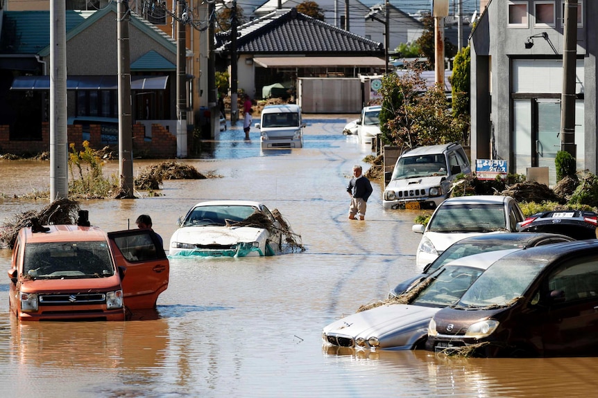 Partially submerged cars are seen in a suburban street, with houses in the background and people walking nearby.