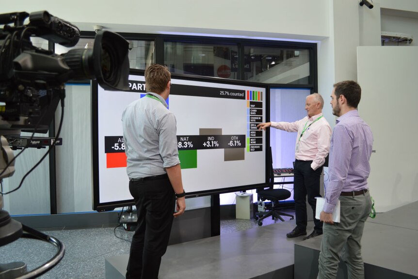 Antony Green standing next to screen and pointing to functions as two men watch on.