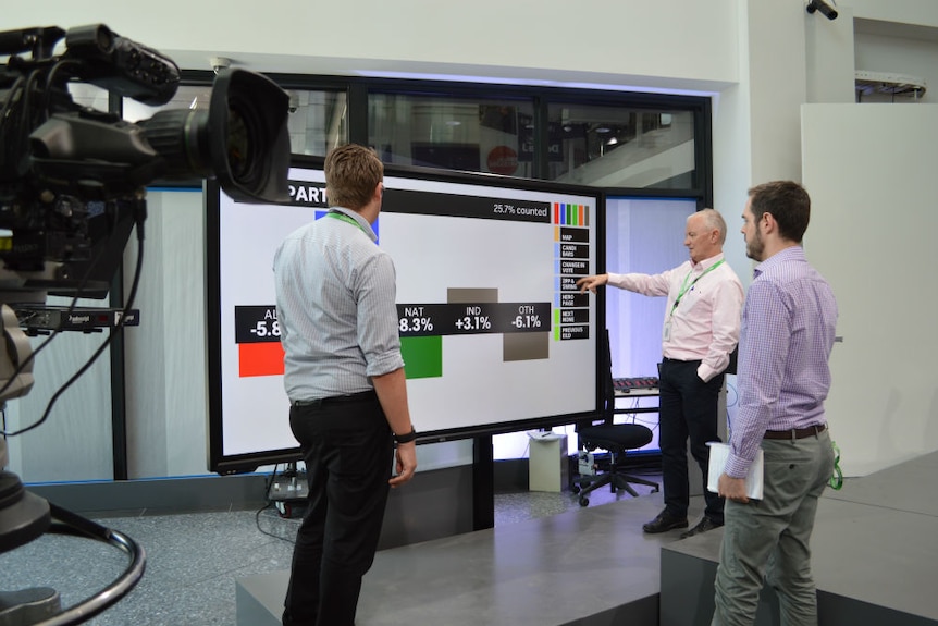 Green pointing to touch screen in foyer of ABC building as two men watch on.