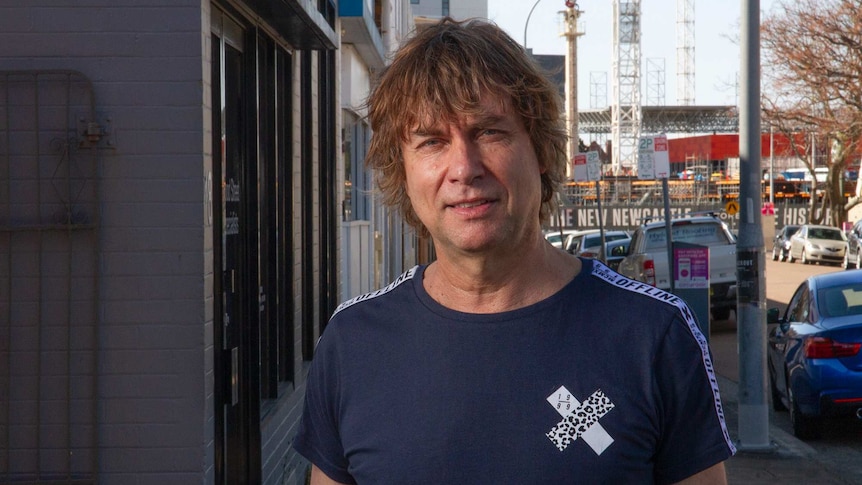 A shaggy haired musician in a blue T-Shirt stands in a street in Newcastle.