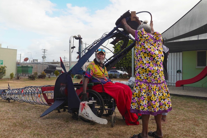 Thaiday in wheelchair metal framed shark sculpture with woman holding mouth open.