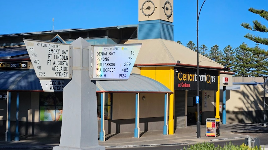 a rainbow shines on street signs in the middle of an intersection with a bottle-o in the background