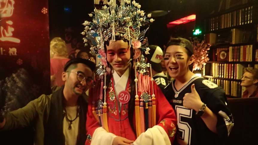 Three people stand together smiling at the camera. In the centre, a drag performer wears colorful robes and a headpiece. 