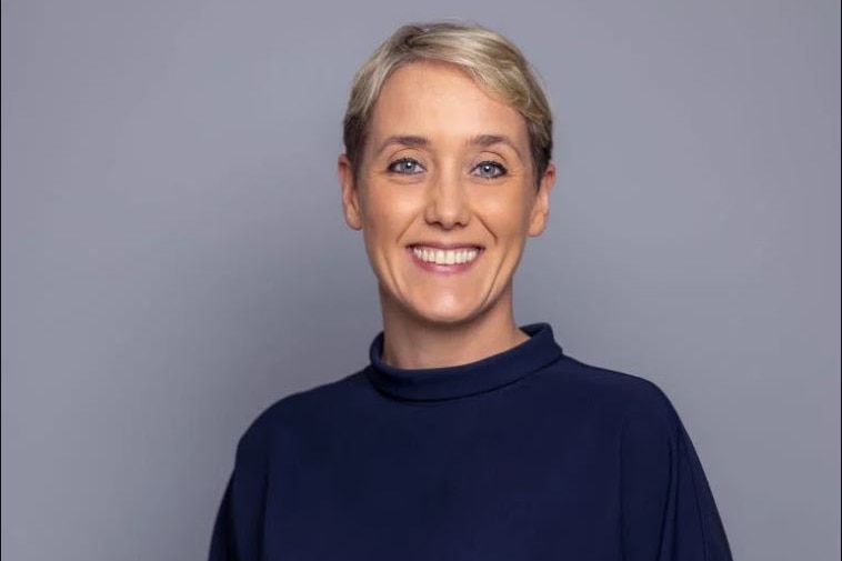 A woman with short blonde hair and a dark blue jumper smiles at a the camera.
