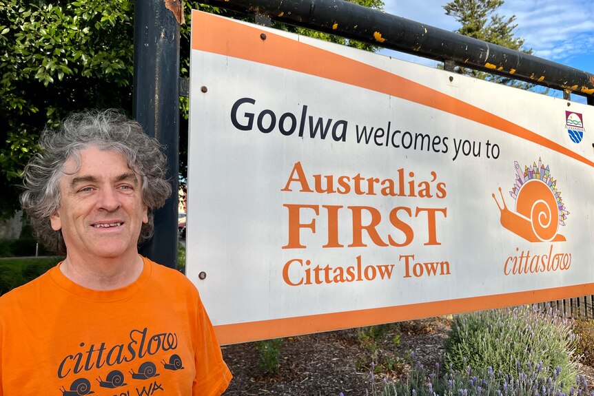 A man in an orange shirt stands next to a sign that declares Goolwa to be Australia's first Cittaslow town