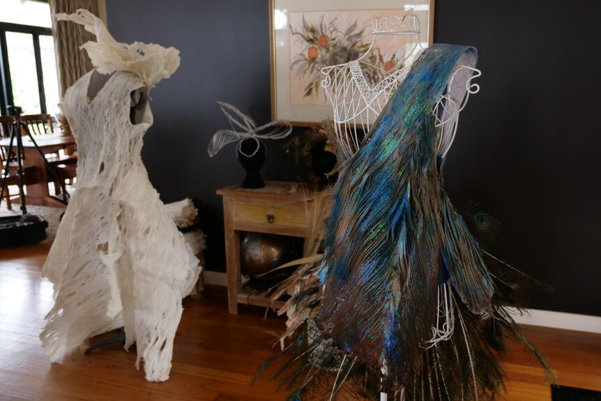 Two dresses, one in cream colours and the other in blue tones, made from plant materials, sit on a display inside a home.